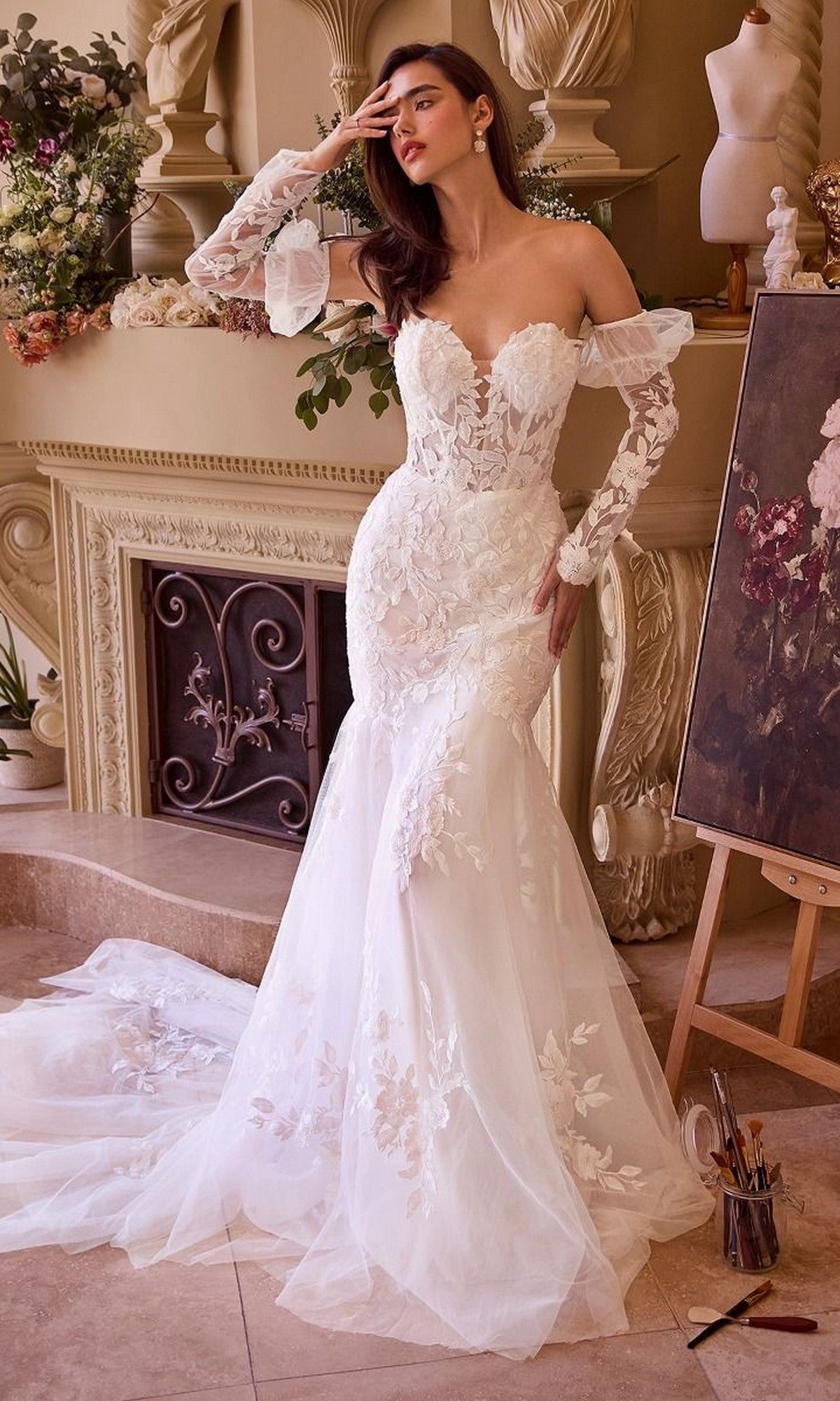 A woman in the Long Wedding Dress: Andrea and Leo WL033, featuring strapless floral lace appliques, stands indoors by a fireplace and an easel with a floral painting.